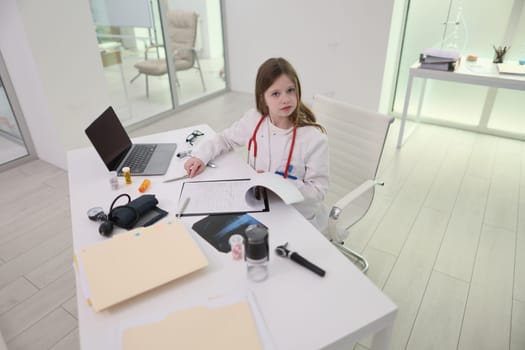 In a chic, minimalist medical office, a young girl dressed as a doctor intently examines a book with great curiosity