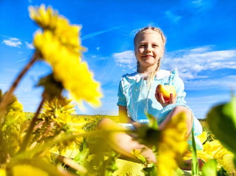 A cheerful girl enjoys a sunny day while sitting among vibrant yellow flowers, holding an apple and smiling at the camera.