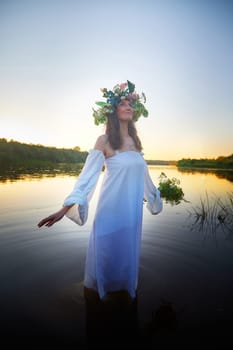 Slavic plump chubby girl in long white dress on the feast of Ivan Kupala with flowers and water in a river or lake on summer evening
