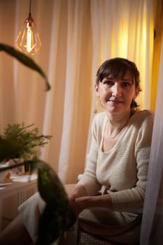 Middle-aged woman sits comfortably in her cozy apartment, enjoying a peaceful evening surrounded by warm lighting and plants.