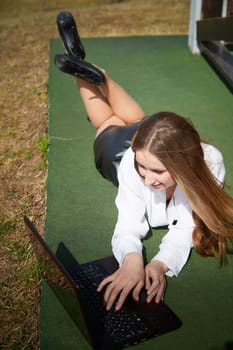 A woman relaxes in the sun, typing on her laptop while lying on her stomach on green grass.