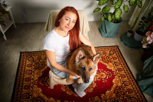 A woman sits comfortably on a chair, gently holding her German Shepherd dog on a patterned rug surrounded by greenery.