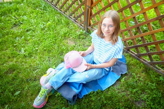 Young girl relaxes on grass in comfortable clothing, holding a pink cap, enjoying a sunny day outdoors near a fence.