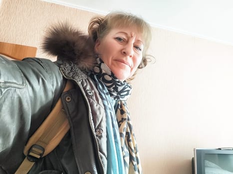 A woman smiles while taking a selfie in a warm winter coat and scarf, standing indoors with a simple backdrop.
