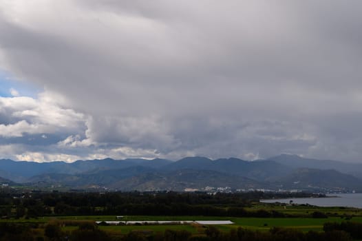 Dramatic clouds billow above vibrant fields while serene water reflects distant mountain silhouettes under gray skies.