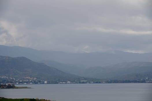 A tranquil lake reflects soft hues as towering mountains linger in the background, shrouded in mist and clouds.