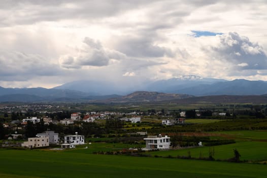 Vast green fields stretch beneath a dramatic sky, framed by distant mountains and modern homes dotting the landscape.