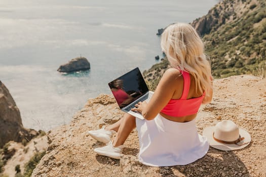 A woman is sitting on a rocky hillside with a laptop open in front of her. She is wearing a pink top and a straw hat. Concept of relaxation and leisure