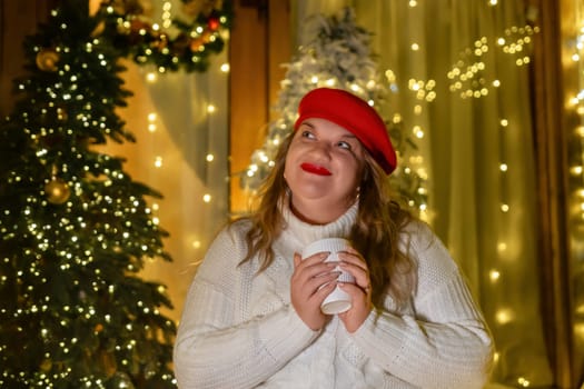 A woman in a white sweater and red beret holds a white mug in front of a Christmas tree with string lights.