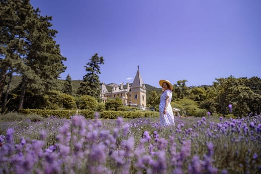 A woman is walking through a field of purple flowers. The field is surrounded by trees and a large house can be seen in the background. The woman is wearing a white dress and a straw hat