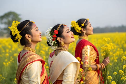 Assamese women in elegant Mekhela Chador, adorned with traditional gold jewelry and fresh flowers in their hair, standing in a yellow mustard field
