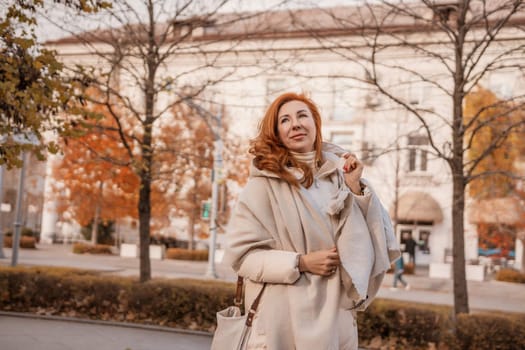 Smiling redhead woman in beige coat stands on autumnal city street, holding a light-colored bag. Fall leaves and a large building are in the background.