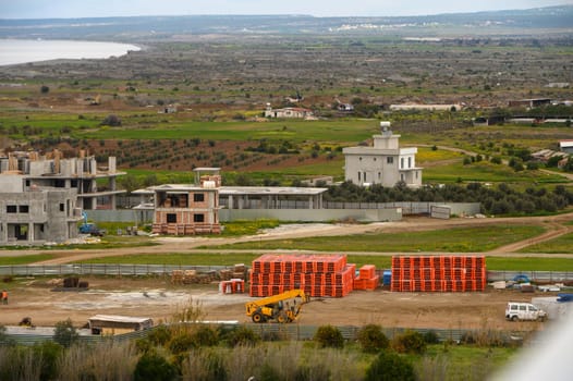 Workers organize materials at a construction site near the coastline, framed by lush green fields and distant hills.