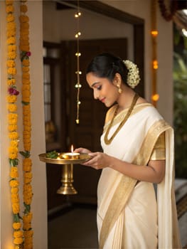 A graceful woman in a white and gold kasavu saree, holding a diya (lamp) with marigold decorations, celebrating a traditional festival.
