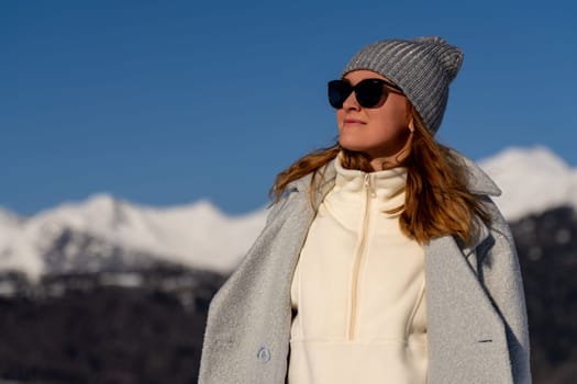 Woman in gray coat and knit hat, sunglasses on, gazes at snow-capped mountains under a clear blue sky. Winter scene.
