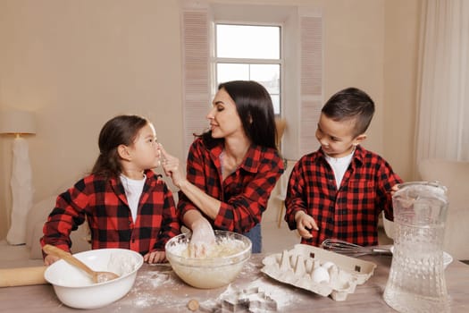 Happy mother and children baking together in a cozy kitchen, wearing matching plaid shirts, playfully touching noses with flour, joyful family bonding moment.
