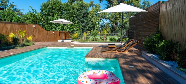 Modern backyard swimming pool with turquoise water, wooden deck, umbrellas, lounge chairs, and pink inflatable ring