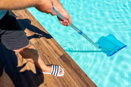 Man cleaning turquoise swimming pool with skimmer net, wooden deck, and sunny outdoor setting