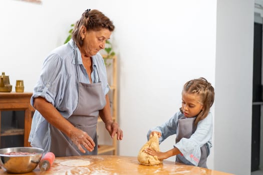 Senior woman lovingly watches her granddaughter knead pastry dough on table, sharing tender and warm moment in comfort of their home
