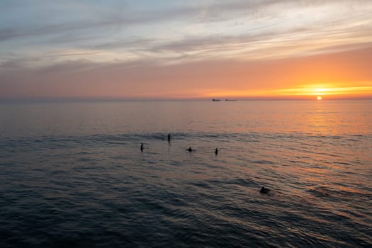 Aerial drone shot of silhouette anonymous people swimming in vast ocean against cloudy sky during sunset. Distant view of tourists amidst seascape enjoying vacation at sundown