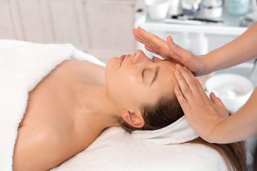 Crop female therapist giving massage on woman's forehead at cosmetology clinic. Relaxed female client is getting pampered in spa by professional beautician
