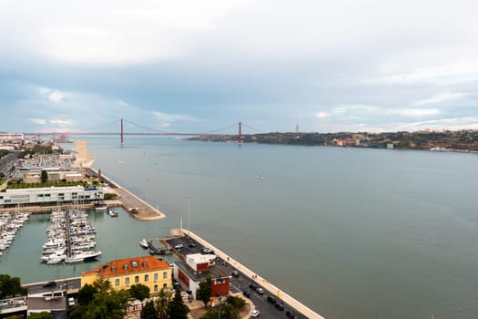 Drone view captures serene harbor by calm waters of Tejo River, set against cloudy sky in Lisbon. The surrounding landscape, with its industrial and urban structures, blends with tranquil waters