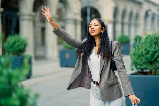 A stylish young woman with beautiful curly hair waves cheerfully while walking confidently through a vibrant urban area, showcasing her impeccable fashion sense and lively personality with every step