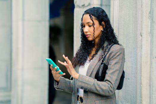 A stylish young woman confidently engages with her smartphone while leaning against a colorful wall in a vibrant urban setting, truly reflecting her modern and contemporary lifestyle