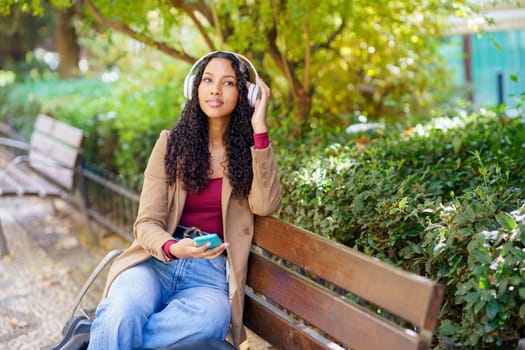 A young woman enjoys her day while sitting on a comfortable bench in a beautiful park. She is wearing stylish headphones, listening to her favorite music, and surrounded by lush, vibrant greenery
