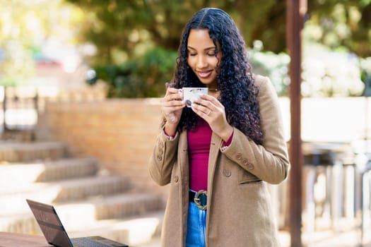 In a serene outdoor workspace, a young woman enjoys sipping her coffee while smiling and engaging with her laptop, surrounded by lush greenery, creating a perfect blend of productivity and relaxation