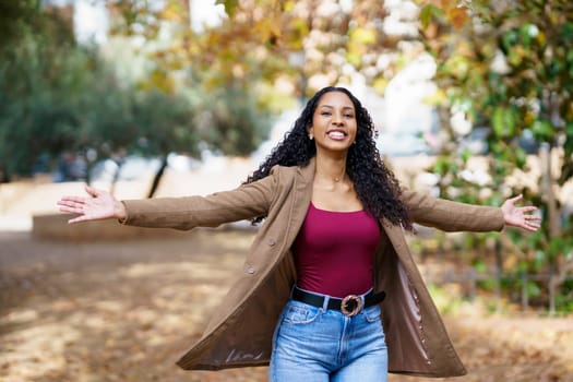 In a sunlit park with lush grass and colorful flowers, a joyful woman with curly hair spreads her arms wide, celebrating the beauty of nature and embracing the moment with freedom