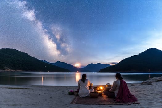 A serene night scene of two people meditating on the beach surrounded by nature, candles, and the Milky Way. A calm and tranquil atmosphere with soft lighting and distant fireworks.