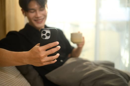 Young man relaxing on a cozy couch, holding coffee cup and using smartphone.