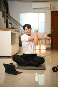Happy asian man stretching his arms while following an online workout on digital tablet.