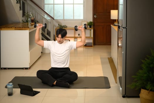 Young man lifting dumbbells while following an online workout on his digital tablet.