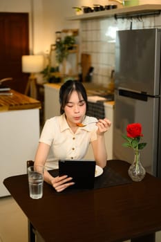 Young woman eating spaghetti and using digital tablet in a decorated home kitchen.