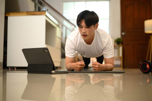 Young man doing plank exercise while following an online workout on digital tablet.