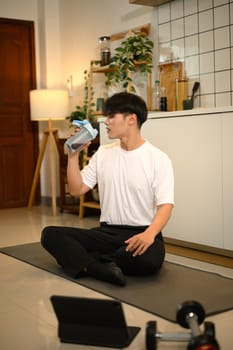 Handsome man sitting on yoga mat, drinking a protein shake after a workout.