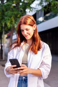 smiling woman using cell phone while walking down the street, she is wearing a white shirt and is focused on his phone, concept of technology of communication and urban lifestyle, copyspace for text