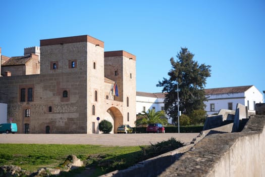 Ancient stone fortress gate with towers and national flags under a clear blue sky in Badajoz, Spain, showcasing its rich history.