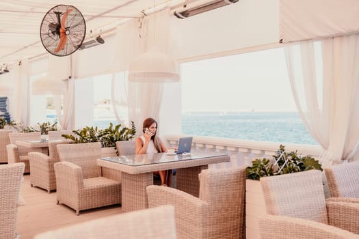 Woman coffee cafe laptop sea. Female freelancer using laptop on coffee break in modern cafe with sea view in the morning.