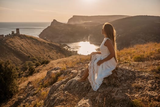 A woman in a white dress sits on a rock overlooking a body of water. The scene is serene and peaceful, with the woman enjoying the view and the calmness of the surroundings