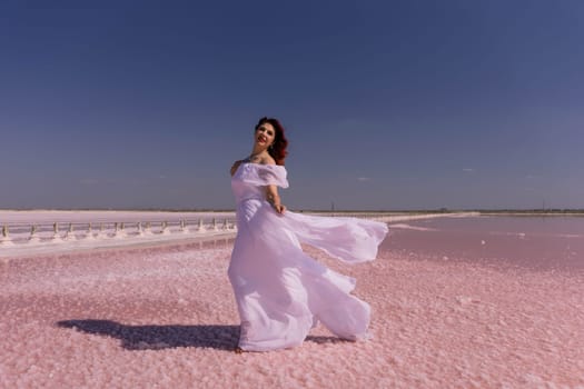 A woman in a white dress stands on a pink salt flat, her dress billowing in the wind, with a clear blue sky behind her.