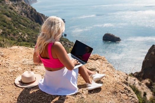 A woman is sitting on a rocky hillside with a laptop open in front of her. She is wearing a pink top and a straw hat. Concept of relaxation and leisure