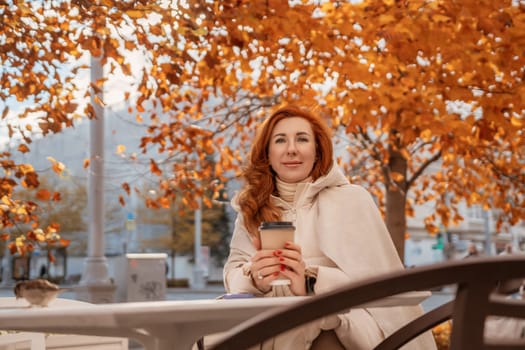 Redhead woman smiles, holding a coffee cup, seated outdoors under vibrant autumn leaves on a park bench.