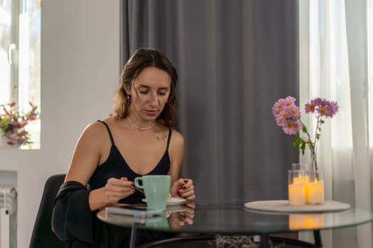 Woman sits at glass table, sipping from teal mug. Dark clothing, flowers and candles nearby. She appears thoughtful.