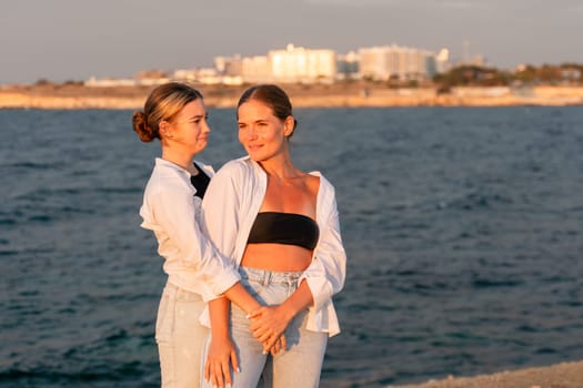 Two women stand close together on a beach, looking at each other. One wears a white shirt and blue jeans, the other a black bandeau top and white shirt. The sea and a city skyline are in the background.