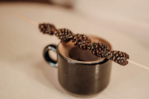 Dark brown coffee in a small mug. A thin skewer with dark brown, spiraled adornments rests across the top. Close-up view on a light beige surface.