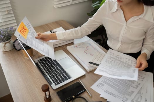A young professional evaluates multiple tax documents while holding a 'TAX' sticky note, highlighting the critical nature of timely tax review in a business environment.