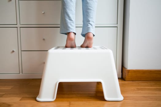 Caucasian child standing on white step stool reaching high cabinet.